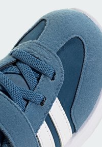 Modrá teniska s textúrovanou tkaninou a semišovými panelmi, biele pruhy po stranách a zladené modré šnúrky viazané do kríža.