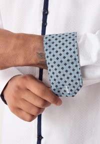 Camicia bianca con motivo a rilievo, polsino geometrico blu con forme esagonali e bottoni blu navy lungo la patta.