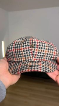 Keps med hundtandsmönster i beige, blå och röda nyanser, tillverkad av texturerat tyg. Har en rundad skärm och två synliga metallöglor.