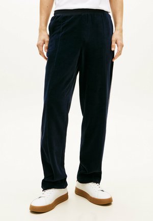 Mand iført mørkeblå fløjlsbukser og hvide sneakers med gummisåler, stående mod en ensfarvet lys baggrund.