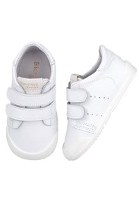 Zapatillas blancas de cuero con cintas de velcro, suela de goma, detalles perforados y logo dorado en la lengüeta. Textura suave, diseño clásico.