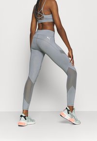 Leggings atletiche grigie con pannelli in rete e logo bianco. Abbinati a un reggiseno sportivo grigio coordinato con un design a incrocio sulla schiena.