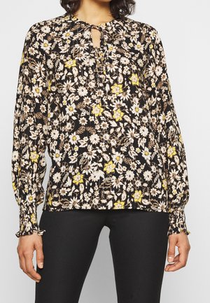 Femme portant un chemisier noir à manches longues avec imprimé floral blanc et jaune et poignets froncés, associé à un pantalon noir.