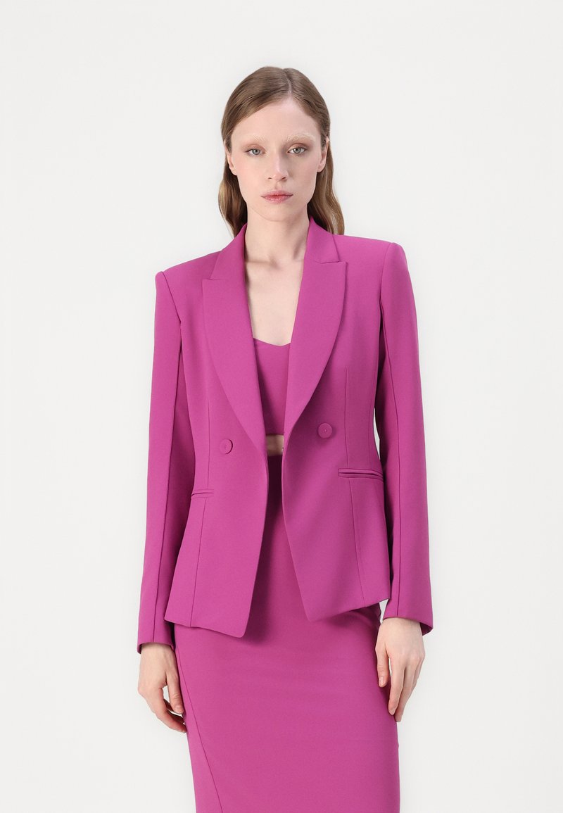 Blazer rose à double boutonnage avec des revers crantés et deux poches avant, associé à une robe rose ajustée. Tissu lisse et design ajusté.