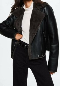 Vrouw in een zwarte leren jasje met grijze bontvoering, witte turtleneck trui, zwarte riem en zwarte broek.