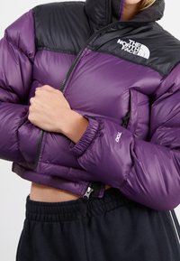 Person wearing a cropped purple and black The North Face puffer jacket with elastic cuffs and a zipper, paired with black sweatpants.