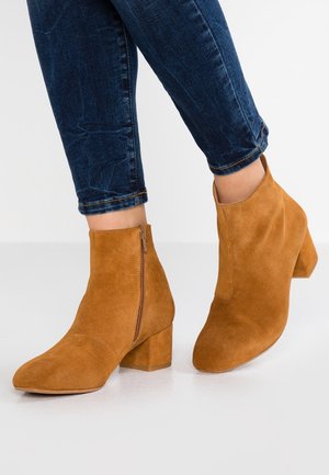 Botas de ante marrón hasta el tobillo con un tacón grueso y cremallera lateral, que presentan una puntera afilada y una textura suave, combinadas con jeans oscuros.