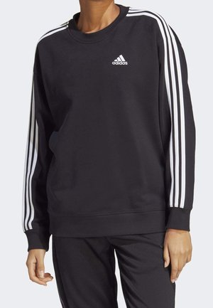 Czarna bluza Adidas z trzema białymi paskami na rękawach i logo na piersi, noszona z dopasowanymi czarnymi spodniami na stojącej osobie.
