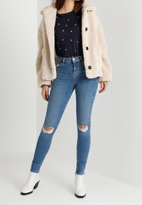 Krémově zbarvená umělá kožešinová bunda s černými knoflíky, v kombinaci s tmavým topem s drobnými, rozptýlenými vzory. Modré džínsy skinny s odřeným vzhledem, bílé kotníčkové boty.