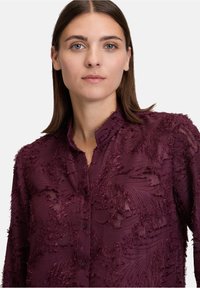 Giovane donna con capelli mossi castani indossa una camicia a bottoni di un viola scuro strutturato con un delicato motivo floreale.