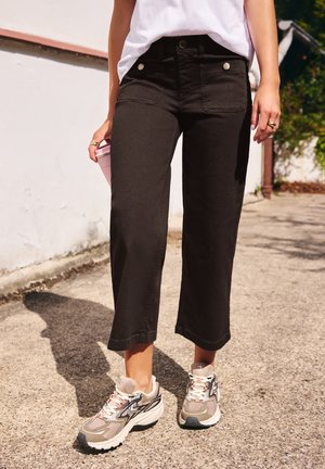 Personne portant un pantalon noir court, des baskets blanches décontractées et une chemise blanche, debout sur un chemin pavé ensoleillé à l'extérieur.