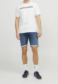 Vit t-shirt med "JACK & JONES"-logotyp, blå denimshorts med viksade fållar, kombinerat med mörka sneakers och vita strumpor.