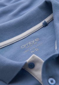 Svetle modré bavlněné polo tričko s bílým límečkem. Obsahuje štítek se značkou "ombre" a velikostí "M". Knoflíkový detail v odpovídající modré barvě.