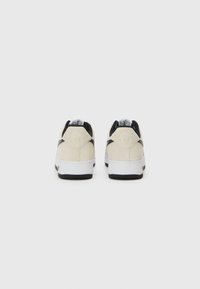 Sneakers with a beige suede upper, black accents, and white rubber sole. Features a distinct heel tab with branding and textured outsole.