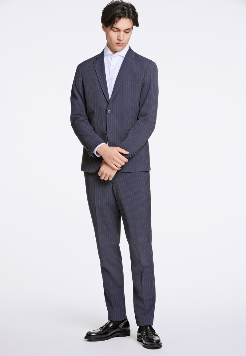 Marinblått pinstripe-kostym med en enknappad blazer och matchande byxor, kombinerat med en vit skjorta och svarta lackskor.