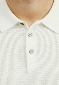 Polo de punto blanco con cuello clásico, tres botones grises y un patrón texturizado. La tela parece ligera y transpirable.