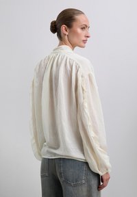 Blusa de crema ligera con mangas con volantes y espalda plisada, combinada con jeans de mezclilla azules. Textura suave y diseño de ajuste relajado.