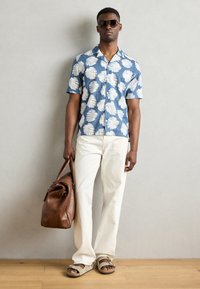 Blauwe shirt met korte mouwen en witte bladerenpatronen, crèmekleurige broek, bruin leren tas en bruine sandalen. Glad materiaal, ontspannen pasvorm.