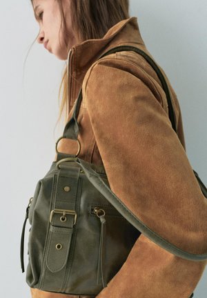 Giovane donna con giacca marrone che porta una borsa a tracolla in pelle verde scuro con fibbia e dettagli con cerniera sulla spalla sinistra.