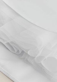 Fehér tüll anyag sima fehér textillel rétegezve; lágy, áttetsző textúrájú, finom fodrokkal a szélén.