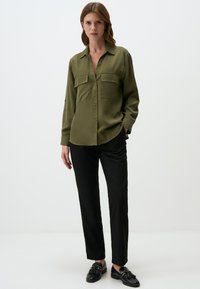 Vrouw in een olijfgroene, knoopsluitende blouse met zakken, zwarte rechte broek en zwarte leren instappers met franjes, staande voor een witte achtergrond.