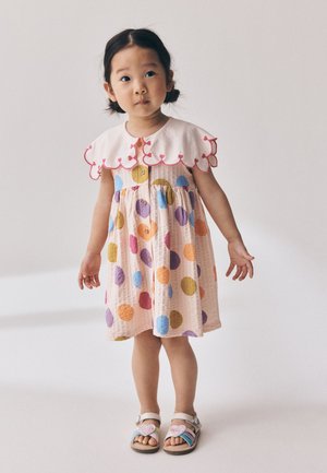 Junges Mädchen mit dunklen Zöpfen, das ein bunt gepunktetes Kleid mit weißem gewellten Kragen und Sandalen trägt, steht vor einem einfarbigen Hintergrund.