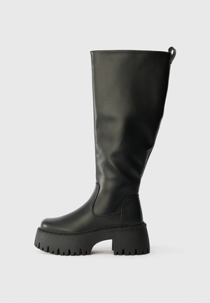 Høye svarte gummistøvler til kneet med grov såle, jevn tekstur og en pull-tab bak for enkel påkledning. Rund tå-design.