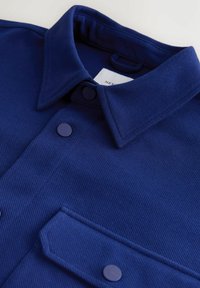 Marineblaues Hemd mit Kragen aus strukturiertem Stoff, mit zwei Knöpfen und einer Brusttasche. Etiketten in der Nähe des Kragens sichtbar.
