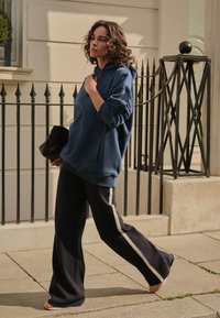 Übergroßer marineblauer Hoodie mit Fronttasche, kombiniert mit marineblauen Hosen mit weitem Bein und einem kontrastierenden cremefarbenen Streifen. Das Modell trägt eine schwarze Tasche.