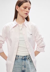 Svijetlo ružičasta košulja s prugama, s dugmićima sprijeda, napravljena od pamuka. Ima mali logo, zavrnute rukave i ležeran kroj. Uparena s bijelom majicom i trapericama visokog struka.