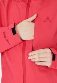 Red waterproof jacket with a high collar, zippered front, and elastic cuffs. Features logo and adjustable cuffs with Velcro.