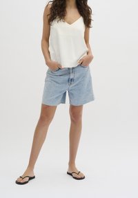 White, sleeveless top with a V-neck, paired with light blue denim shorts, featuring a raw hem and loose fit. Black flip-flops complete the look.