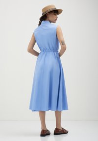 Abito blu senza maniche con design da camicia, vita elastica e gonna fluida. Abbinato a un cappello di paglia e scarpe marroni.
