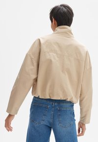 Beige cropped Jacke mit hohem Kragen, mit einer glatten Textur und tief sitzenden Schultern, getragen über blauen Jeans mit Gesäßtaschen.