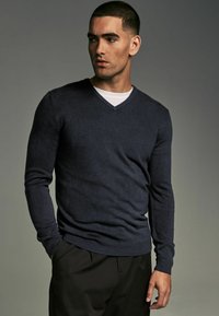 Pulover bleumarin cu decolteu în V, realizat dintr-un material moale, cu mâneci lungi și un design ajustat. Asortat cu un tricou alb cu guler rotund dedesubt.