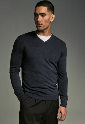 Navy v-neck sweater made of soft fabric, featuring long sleeves and a fitted design. Paired with a white crew neck shirt underneath.