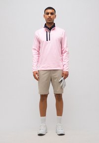 Mężczyzna w różowym pulowerze na zamek do ćwiartki, beżowych szortach, białych trampkach, trzymający w prawej ręce pojedynczą szarą rękawiczkę golfową.