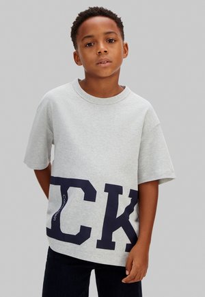 Mladý chlapec s krátkými kudrnatými vlasy, oblečený ve šedém volném tričku s velkými tmavě modrými písmeny a černých kalhotách na jednobarevném pozadí.