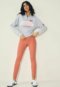 Šedá mikina s potiskem "BOSTON" v červené barvě, se zipovým límcem, spárovaná s korálovými slim-fit kalhotami a černobílými teniskami. Hladký texture.