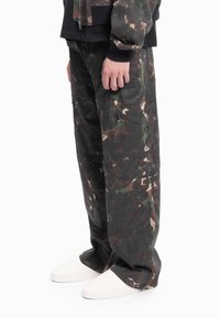 Kamuflasjebukser i mørkegrønne og brune toner, med cargolommer, løs passform og kontrasterende hvite sko. Teksturert stoff.