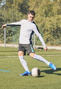Conjunto deportivo de Nike: camiseta de manga larga blanca con detalles en negro, pantalones cortos grises, calcetas blancas y zapatillas de fútbol azules. Pateando un balón de fútbol sobre la hierba.