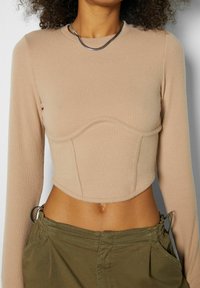 Femme portant un crop top à manches longues côtelé beige avec des détails de couture et un pantalon vert olive, accessoirisée d'un collier chaîne argenté.