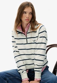 Wit sweatshirt met marineblauwe horizontale strepen, hoge kraag en een rits aan de voorkant. Daaronder is een roze geruit overhemd zichtbaar.