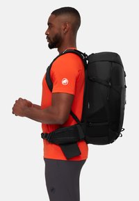Sac à dos noir avec des bretelles ergonomiques, des poches en maille sur les côtés et une poignée supérieure, porté par une personne en chemise orange et leggings foncés.