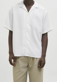 Uomo che indossa una camicia bianca a maniche corte con sottili righe e pantaloni beige su uno sfondo semplice.