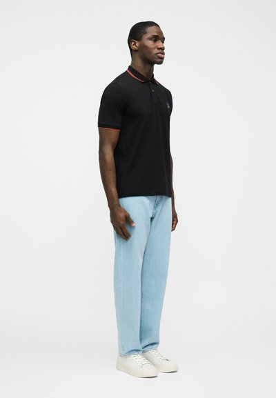 Άνδρας που στέκεται πλαγίως, φοράει μαύρο πόλο μπλουζάκι με κόκκινη πατίτα, ανοιχτό μπλε τζιν και λευκά αθλητικά παπούτσια, μπροστά σε απλό λευκό φόντο.