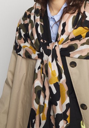 Femme portant un trench-coat beige et une chemise bleue à col, avec une grande écharpe à motif abstrait multicolore drapée sur les épaules.