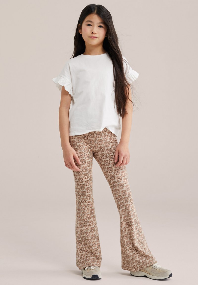Fille aux longs cheveux bruns portant un haut blanc à manches volantées, un pantalon évasé beige à motifs floraux et des baskets claires, debout.