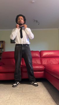 Chemise blanche à manches longues et cravate noire associée à un jean foncé et des baskets noires. Se tenant devant un canapé rouge, en train d'ajuster la cravate.