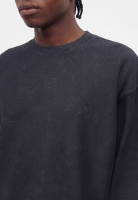 Човек, носещ черен суитшърт Carhartt с кръгло деколте и видимо бродирано лого на марката от лявата страна на гърдите.